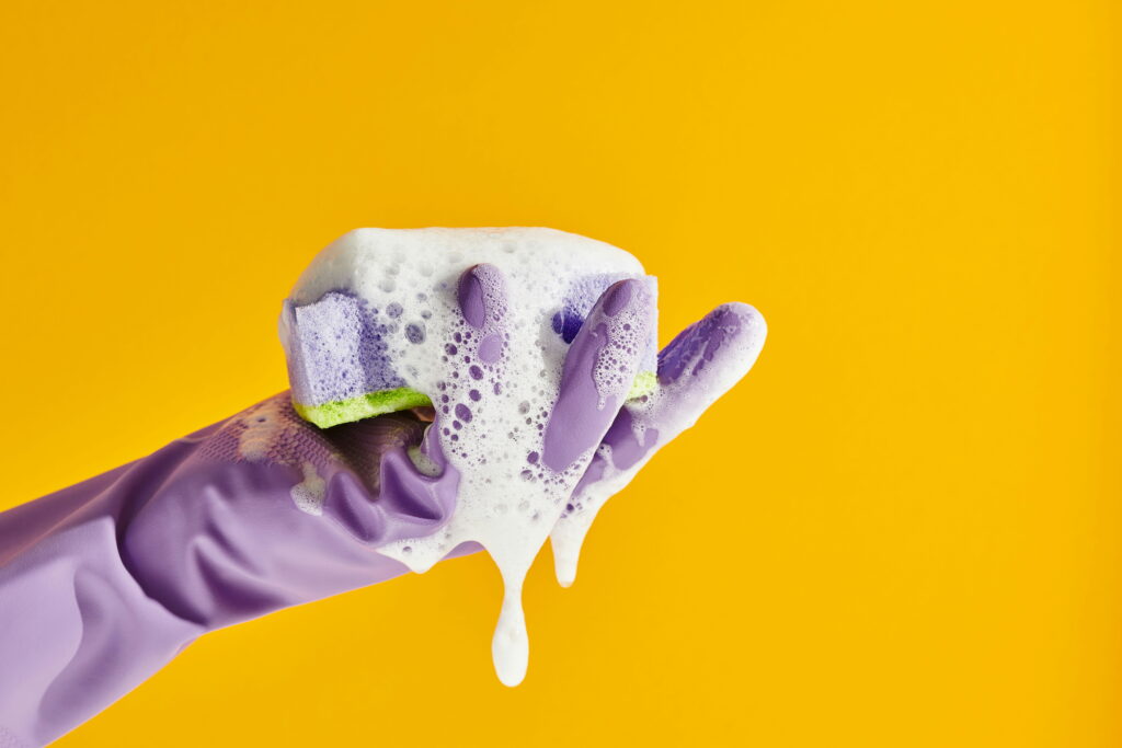 Gloved hand squeezing a bubbly purple sponge with white foam against a bright yellow background.